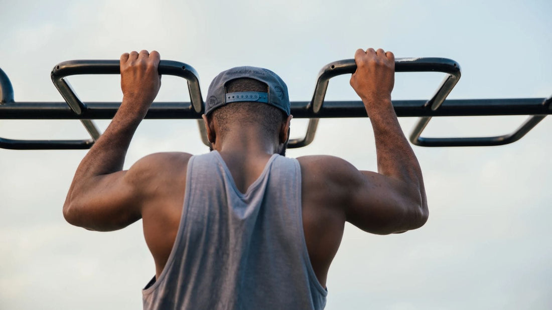 Man with grey muscle shirt and hat performing pull ups on a black bar 